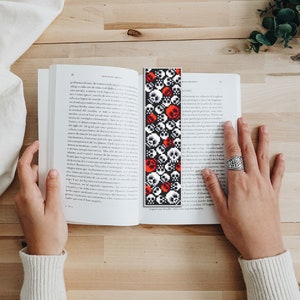 May include: A black and white bookmark with a repeating pattern of red and white skulls. The bookmark is being held in a hand with a silver ring.