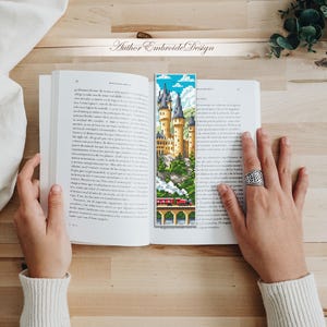 Puede incluir: Un marcap&aacute;ginas colorido con una imagen pixelada de un castillo, un tren y un cielo azul con nubes. El marcap&aacute;ginas est&aacute; colocado en un libro abierto. El texto "Author Embroide Design" es visible.