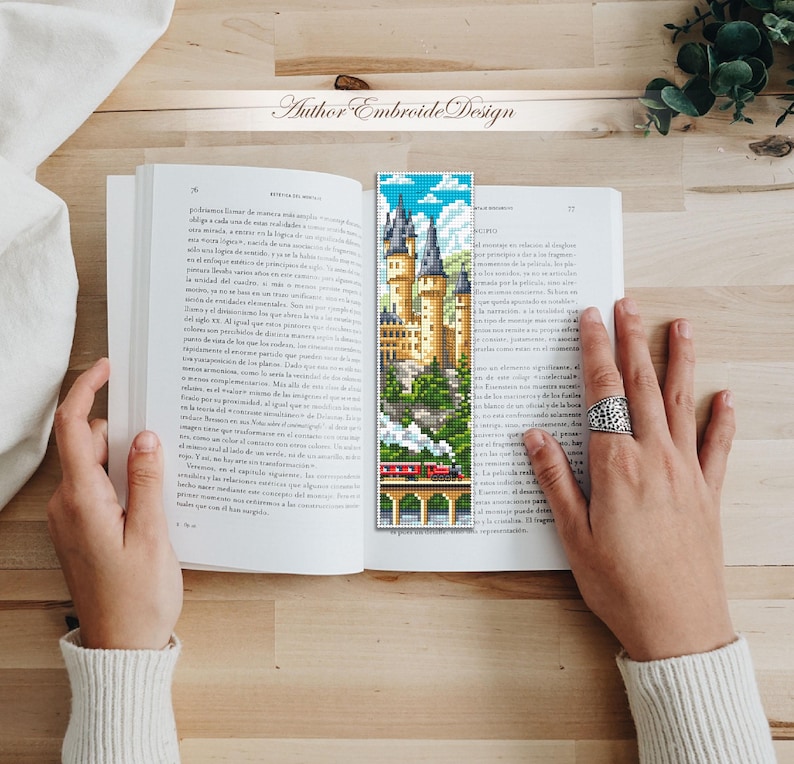 Puede incluir: Un marcap&aacute;ginas colorido con una imagen pixelada de un castillo, un tren y un cielo azul con nubes. El marcap&aacute;ginas est&aacute; colocado en un libro abierto. El texto "Author Embroide Design" es visible.