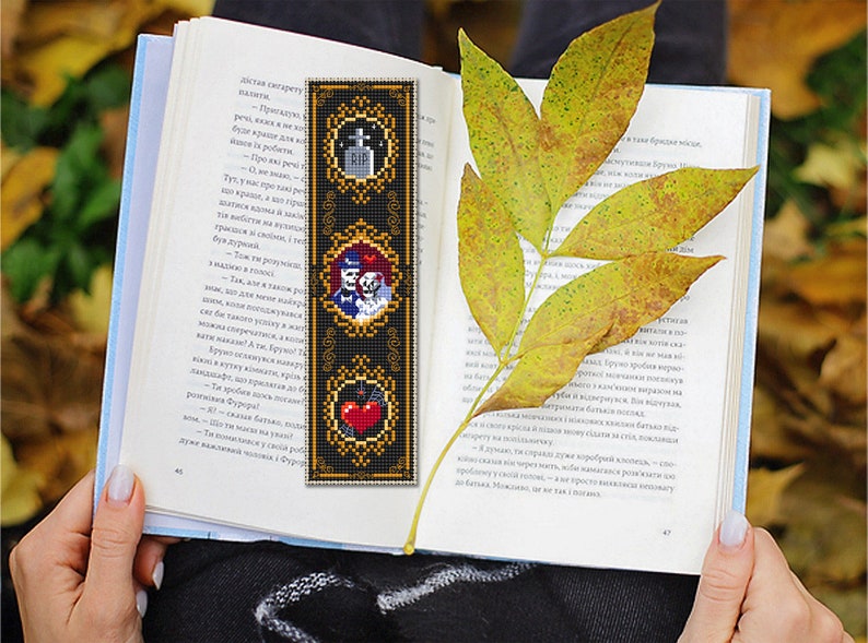 May include: A black bookmark with gold trim features three framed images. The top image shows a tombstone with the letters RIP. The middle image shows a skeleton couple in a wedding pose. The bottom image shows a red heart with a bat.