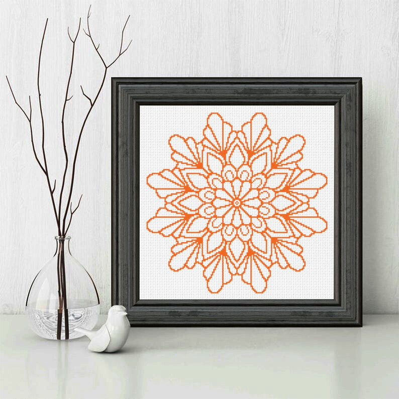 May include: A framed cross-stitch pattern of an orange mandala design. The mandala is a circular design with intricate details and a floral motif.