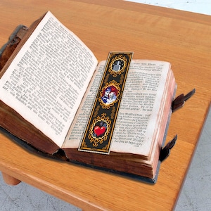 May include: A black and gold bookmark with a red heart, a skull and crossbones, and a pair of theater masks. The bookmark is placed in an open antique book.
