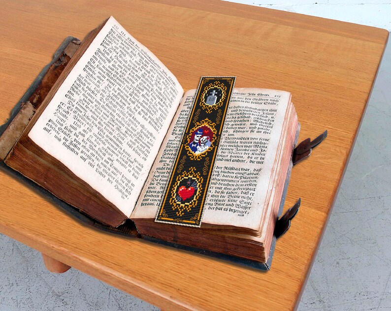 May include: A black and gold bookmark with a red heart, a skull and crossbones, and a pair of theater masks. The bookmark is placed in an open antique book.