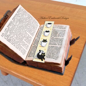 Puede incluir: Un marcap&aacute;ginas con un dise&ntilde;o de gato negro. El gato est&aacute; sentado en una taza de t&eacute; con una taza de t&eacute; humeante. El marcap&aacute;ginas es amarillo con contornos negros y el texto "Author Embroide Design".