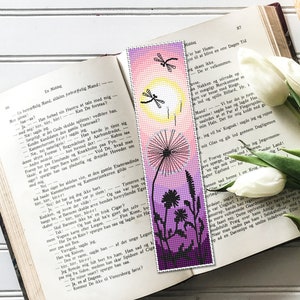 May include: A handmade bookmark featuring a dandelion design with a gradient background of pink, yellow, and purple. The bookmark has a beaded appearance and includes dragonfly silhouettes. It rests on an open book with text.