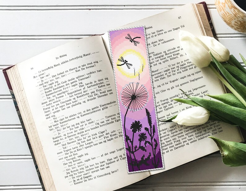 May include: A handmade bookmark featuring a dandelion design with a gradient background of pink, yellow, and purple. The bookmark has a beaded appearance and includes dragonfly silhouettes. It rests on an open book with text.