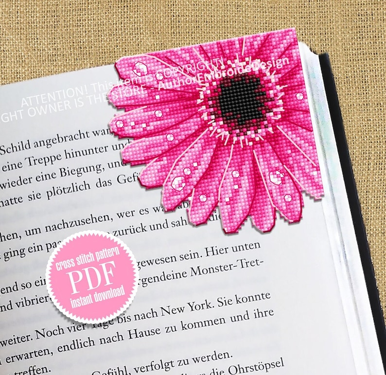 Puede incluir: Un marcap&aacute;ginas rosa de punto de cruz con forma de gerbera, con detalles de gotas de agua. El marcap&aacute;ginas est&aacute; colocado en la esquina de un libro. Tambi&eacute;n es visible un c&iacute;rculo rosa con el texto "cross stitch pattern PDF instant download".