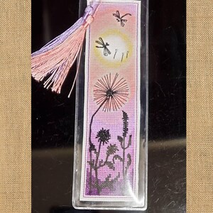 May include: Embroidered bookmark featuring a dandelion, dragonflies, and a sunset design. The bookmark has a pink and purple tassel. The text on the bookmark says "Embroidered bookmark by our customer Cindy Kling."