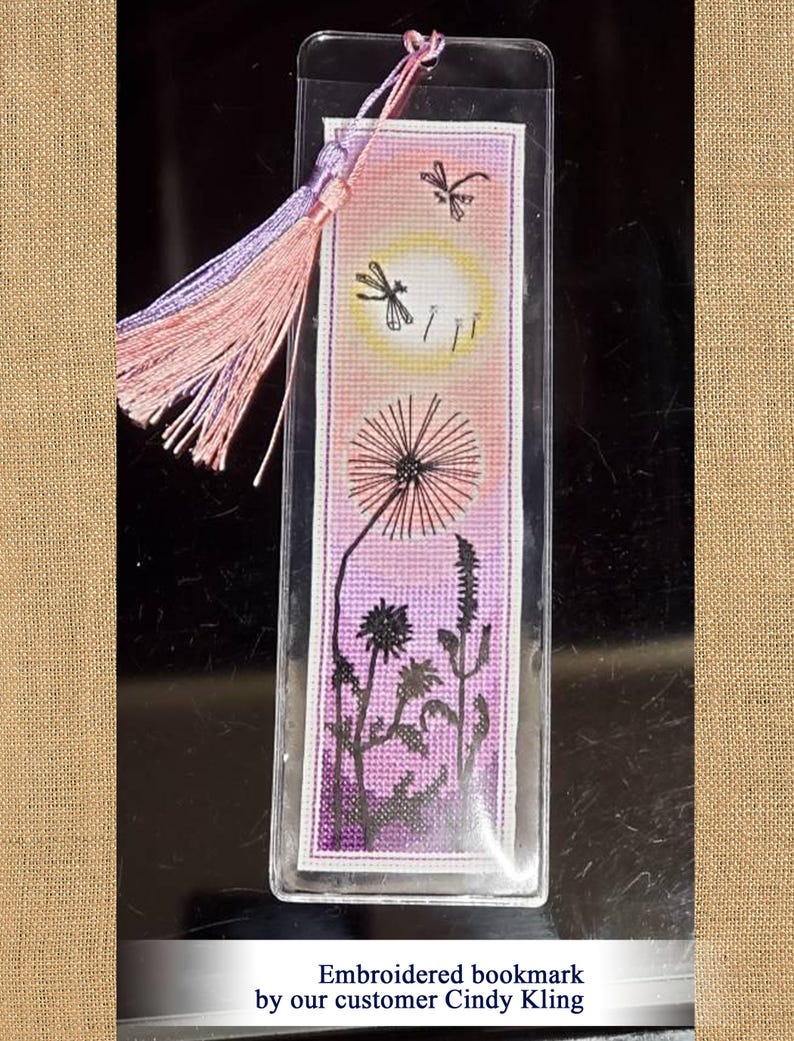 May include: Embroidered bookmark featuring a dandelion, dragonflies, and a sunset design. The bookmark has a pink and purple tassel. The text on the bookmark says "Embroidered bookmark by our customer Cindy Kling."