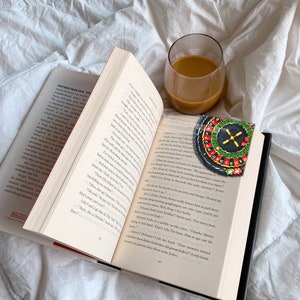 Op de afbeelding: Een close-up van een open boek met een glas koffie en een kralenroulette-bladwijzer. Het boek is open op een pagina met tekst over een tienermeisje en haar vrienden. De roulette-bladwijzer is rood, zwart en groen met witte nummers.