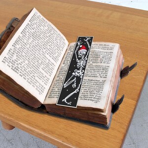 May include: A black and white bookmark with a skeleton wearing a red Santa hat. The bookmark is placed in an open book with German text.