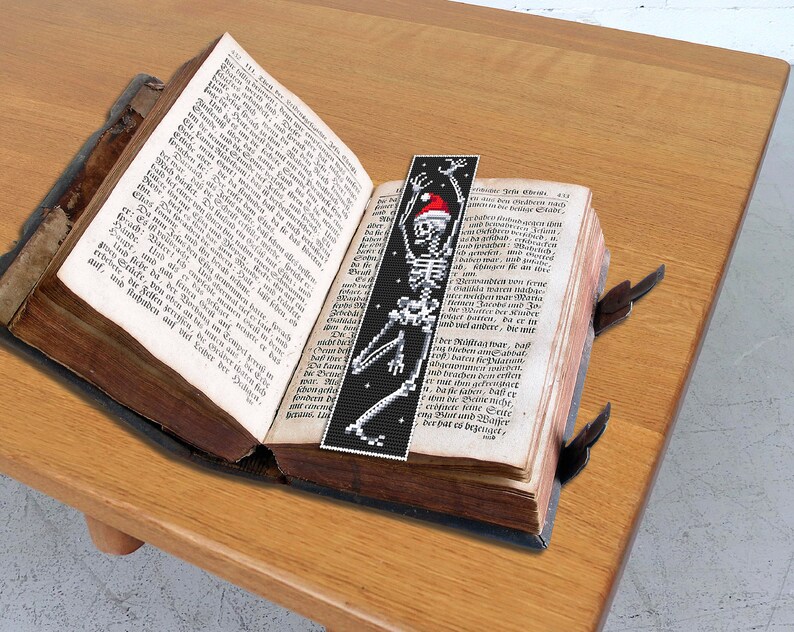 May include: A black and white bookmark with a skeleton wearing a red Santa hat. The bookmark is placed in an open book with German text.