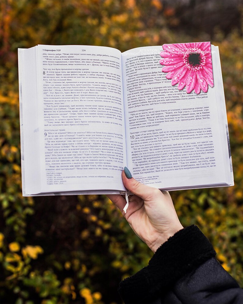Puede incluir: Un libro abierto con texto, sostenido por una persona. Un marcap&aacute;ginas en forma de margarita rosa se inserta entre las p&aacute;ginas. El fondo est&aacute; borroso con follaje verde y amarillo.