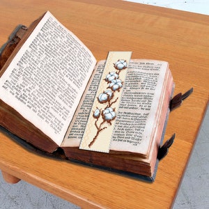 May include: A close-up of a vintage book with a bookmark featuring a white cotton flower design. The bookmark is made of fabric and has a brown background. The book is open to a page with German text.