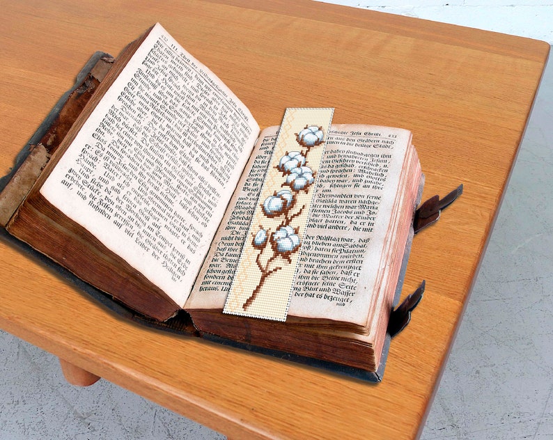 May include: A close-up of a vintage book with a bookmark featuring a white cotton flower design. The bookmark is made of fabric and has a brown background. The book is open to a page with German text.