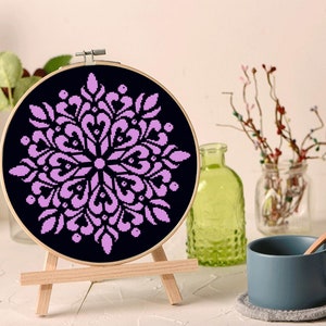 May include: A round, black cross-stitch embroidery hoop featuring a symmetrical, lavender floral design. The embroidery is displayed on a small wooden easel, with a blue mug and decorative bottles in the background.