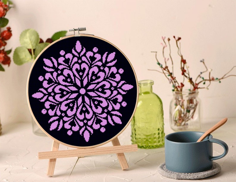 May include: A round, black cross-stitch embroidery hoop featuring a symmetrical, lavender floral design. The embroidery is displayed on a small wooden easel, with a blue mug and decorative bottles in the background.