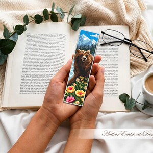 May include: A handmade bookmark featuring a detailed cross-stitch design of a brown bear in a mountain landscape with colorful flowers. The bookmark is held over an open book, with a cup of coffee and glasses nearby.