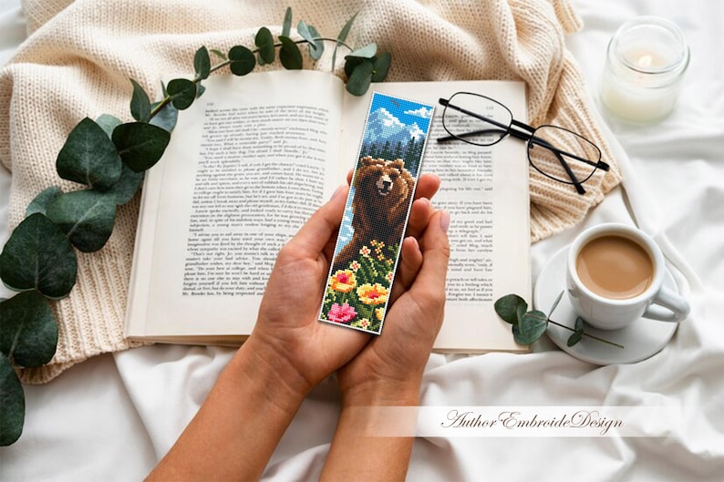 May include: A handmade bookmark featuring a detailed cross-stitch design of a brown bear in a mountain landscape with colorful flowers. The bookmark is held over an open book, with a cup of coffee and glasses nearby.