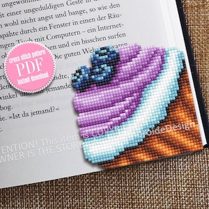 Puede incluir: Un patrón de punto de cruz de un cupcake morado y azul con glaseado blanco y arándanos en la parte superior. El cupcake está sentado sobre una base marrón. El patrón está disponible para descargar en PDF.