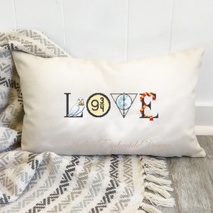 May include: A white pillow with the word "LOVE" embroidered on it. Each letter is decorated with a different Harry Potter themed icon: a snowy owl, the platform 9 3/4 sign, the Deathly Hallows symbol, and a red and gold scarf.