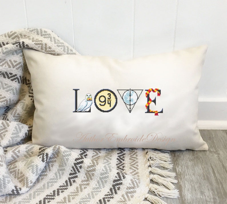 May include: A white pillow with the word "LOVE" embroidered on it. Each letter is decorated with a different Harry Potter themed icon: a snowy owl, the platform 9 3/4 sign, the Deathly Hallows symbol, and a red and gold scarf.
