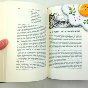May include: Open book with a green bookmark featuring a yellow and white embroidered image of a fried egg. The book's pages are filled with text, including the title "The Two Towers" and the chapter title "Of Herbs and Stewed Rabbit".