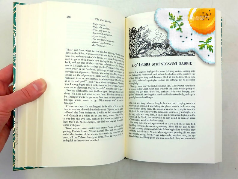 May include: Open book with a green bookmark featuring a yellow and white embroidered image of a fried egg. The book's pages are filled with text, including the title "The Two Towers" and the chapter title "Of Herbs and Stewed Rabbit".