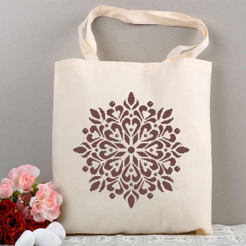 May include: Cream-colored tote bag with a brown, symmetrical floral design. The bag has matching cream-colored handles and is made of a sturdy fabric. The floral design is centered on the front of the bag. The bag is sitting next to a bouquet of flowers.