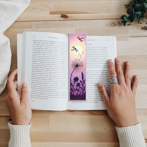 May include: A handmade bookmark featuring a pixelated design of a dandelion, dragonflies, and a sun against a gradient of pink, purple, and yellow. The bookmark is placed in an open book.