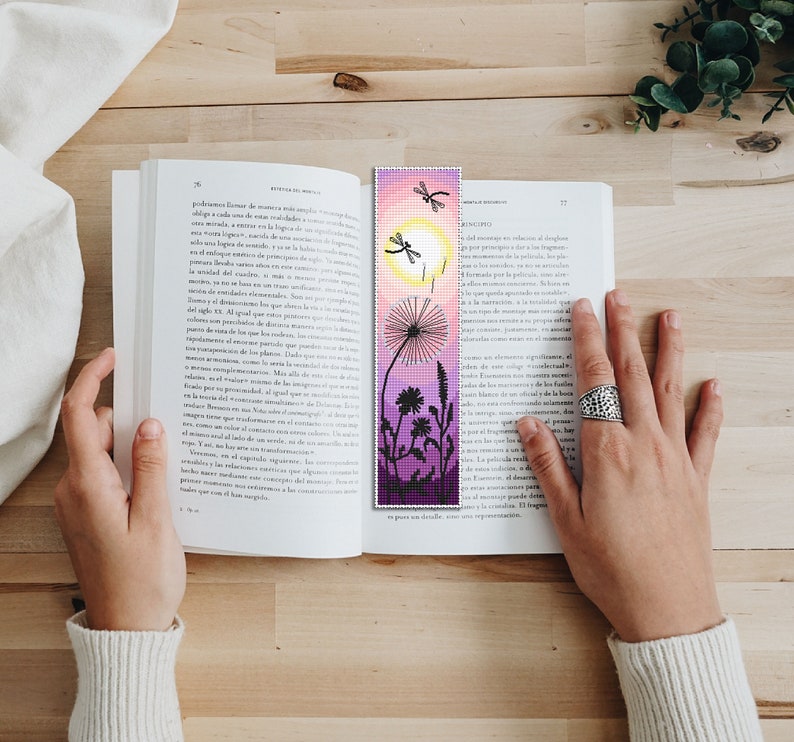 May include: A handmade bookmark featuring a pixelated design of a dandelion, dragonflies, and a sun against a gradient of pink, purple, and yellow. The bookmark is placed in an open book.