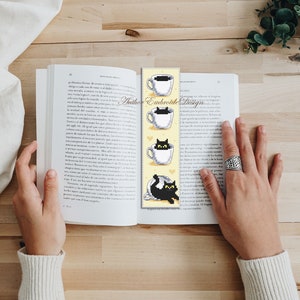 Puede incluir: Un marcap&aacute;ginas amarillo con un gato negro asom&aacute;ndose a una taza de t&eacute; blanca. El marcap&aacute;ginas tiene el texto "Author Embroide Design" impreso en &eacute;l.