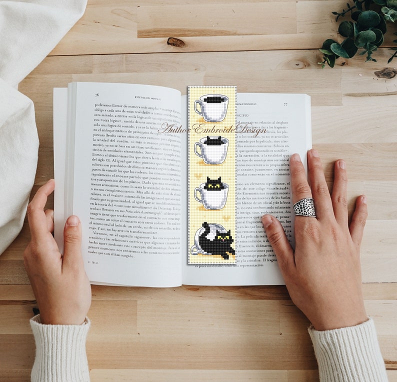 Puede incluir: Un marcap&aacute;ginas amarillo con un gato negro asom&aacute;ndose a una taza de t&eacute; blanca. El marcap&aacute;ginas tiene el texto "Author Embroide Design" impreso en &eacute;l.