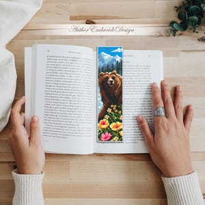 May include: A handmade bookmark featuring a pixelated image of a brown bear with a mountain backdrop and colorful flowers. The bookmark is placed in an open book. The text "Author Embroide Design" is visible.