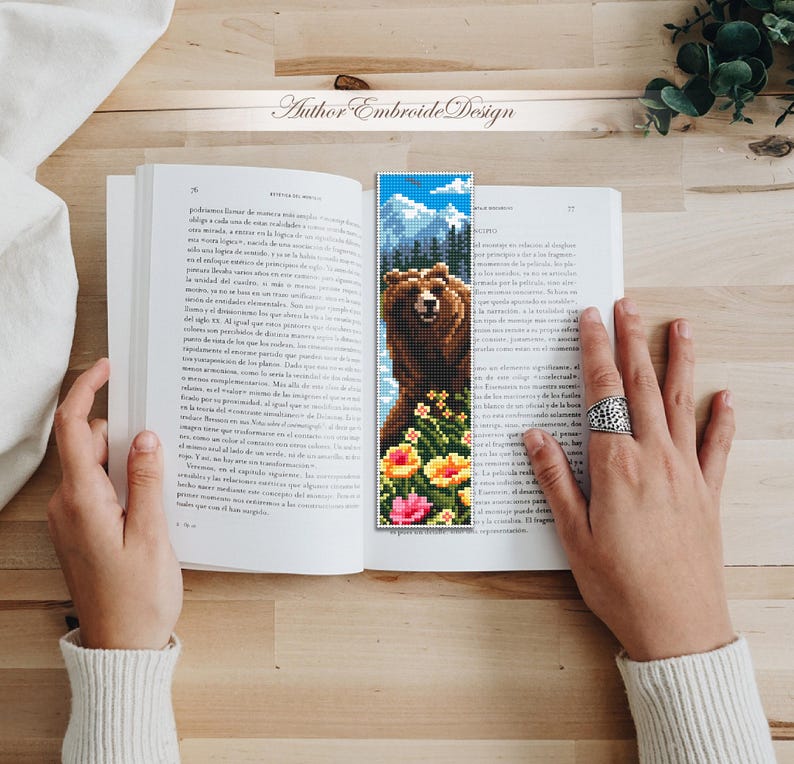 May include: A handmade bookmark featuring a pixelated image of a brown bear with a mountain backdrop and colorful flowers. The bookmark is placed in an open book. The text "Author Embroide Design" is visible.