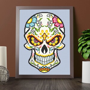 Puede incluir: Una impresi&oacute;n enmarcada de una calavera de az&uacute;car colorida con dise&ntilde;os florales y una cruz en la frente. La calavera es blanca con contornos negros y presenta flores amarillas, rosas, azules y verdes.