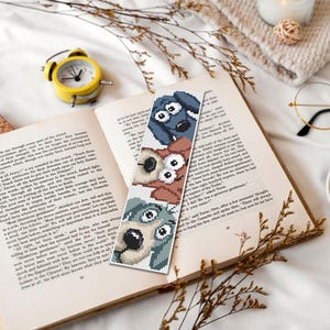 May include: A handmade bookmark with three cartoon dog faces in blue, brown, and green. The bookmark is in an open book, with a yellow alarm clock, glasses, and a cup of tea. The scene is on a white surface.