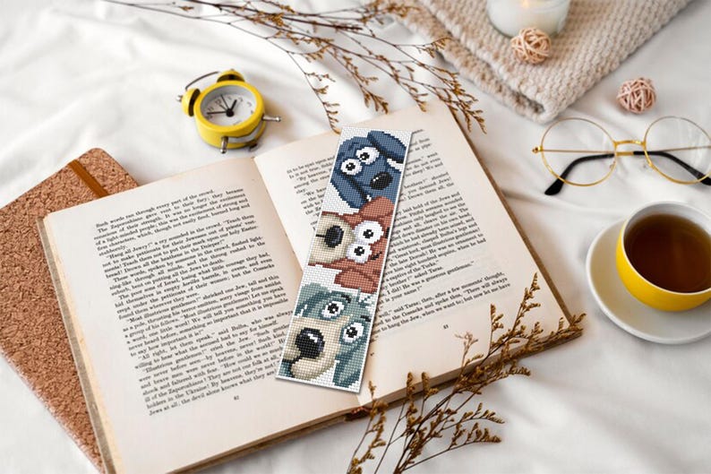 May include: A handmade bookmark with three cartoon dog faces in blue, brown, and green. The bookmark is in an open book, with a yellow alarm clock, glasses, and a cup of tea. The scene is on a white surface.