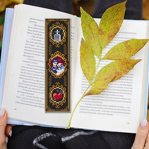 May include: A black bookmark with gold trim features three framed images. The top image shows a tombstone with the letters RIP. The middle image shows a skeleton couple in a wedding pose. The bottom image shows a red heart with a bat.