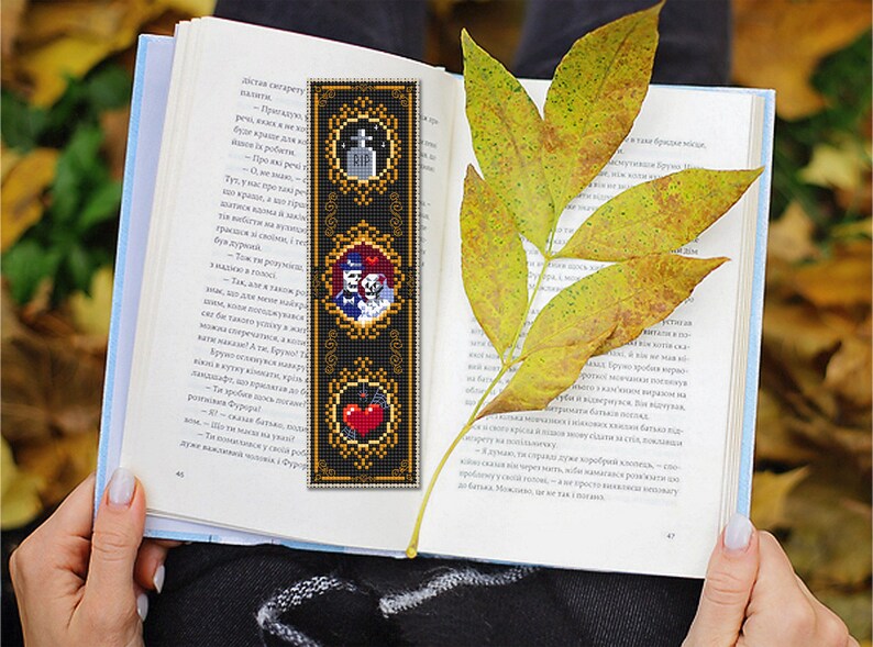 May include: A black bookmark with gold trim features three framed images. The top image shows a tombstone with the letters RIP. The middle image shows a skeleton couple in a wedding pose. The bottom image shows a red heart with a bat.