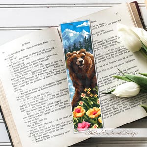 May include: A handmade beaded bookmark featuring a brown bear, flowers, and a mountain scene. The bookmark is placed on an open book with white tulips nearby. The bookmark measures approximately 2 x 7 inches.