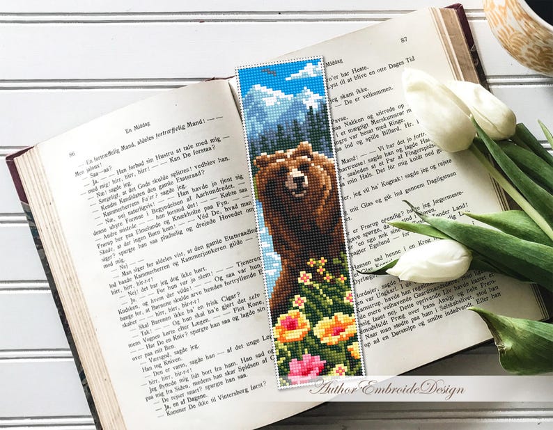 May include: A handmade beaded bookmark featuring a brown bear, flowers, and a mountain scene. The bookmark is placed on an open book with white tulips nearby. The bookmark measures approximately 2 x 7 inches.