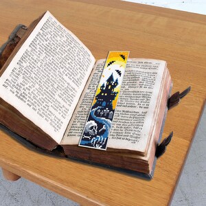 Puede incluir: Un marcap&aacute;ginas azul y amarillo con una silueta negra de una casa encantada y murci&eacute;lagos. El marcap&aacute;ginas est&aacute; colocado en un libro abierto.