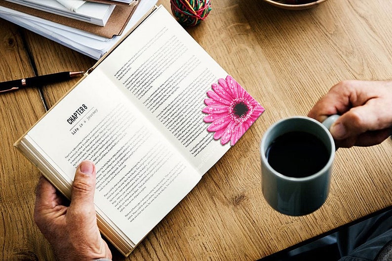 Puede incluir: Un libro abierto con el Cap&iacute;tulo 8 titulado "La vida es un viaje" est&aacute; sobre una mesa de madera. Un marcap&aacute;ginas de gerbera rosa con gotas de agua descansa en la p&aacute;gina. Una taza gris llena de l&iacute;quido oscuro est&aacute; cerca.
