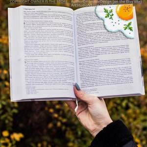 May include: A close-up of a hand holding open a book with a bookmark featuring a pixelated image of a fried egg with green parsley. The book is open to a page with text in a foreign language. The background is blurred and shows a fall foliage scene.