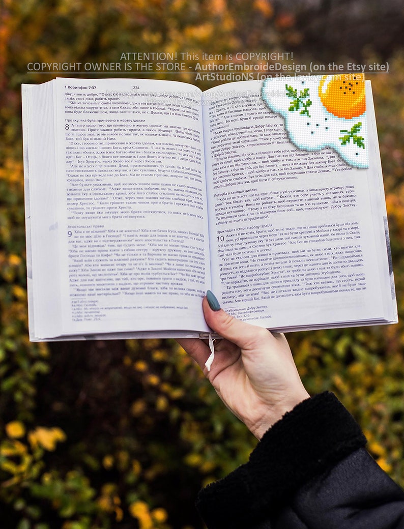 May include: A close-up of a hand holding open a book with a bookmark featuring a pixelated image of a fried egg with green parsley. The book is open to a page with text in a foreign language. The background is blurred and shows a fall foliage scene.