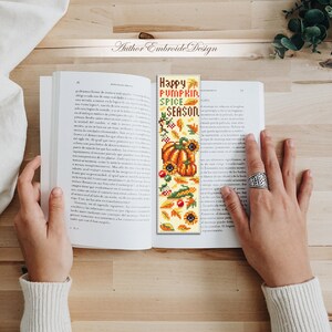 May include: A handmade bookmark with the text "Happy Pumpkin Spice Season" and fall-themed illustrations. The bookmark features pumpkins, sunflowers, leaves, and berries in shades of orange, yellow, and green. The bookmark is held open in a book.