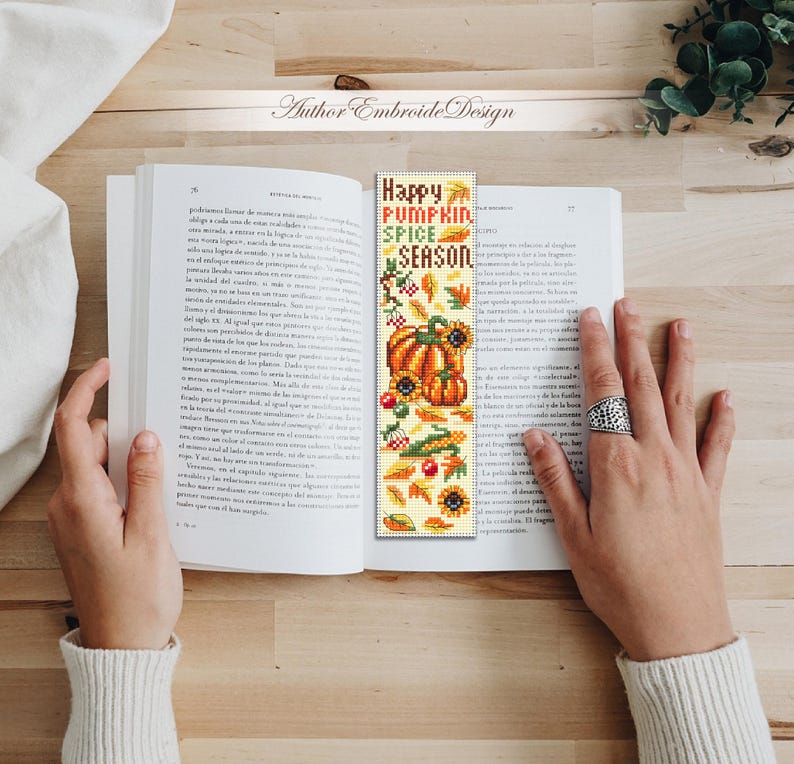 May include: A handmade bookmark with the text "Happy Pumpkin Spice Season" and fall-themed illustrations. The bookmark features pumpkins, sunflowers, leaves, and berries in shades of orange, yellow, and green. The bookmark is held open in a book.