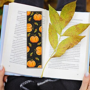 May include: A black bookmark with a repeating pattern of orange pumpkins and green leaves. The bookmark is made of fabric and has a fringed edge.