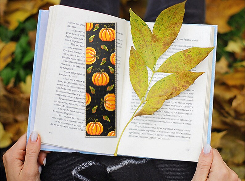 May include: A black bookmark with a repeating pattern of orange pumpkins and green leaves. The bookmark is made of fabric and has a fringed edge.
